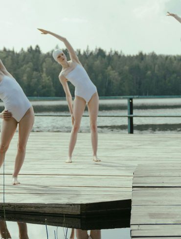 Three outdoor swimmers in white swimming costumes stretch by a lake