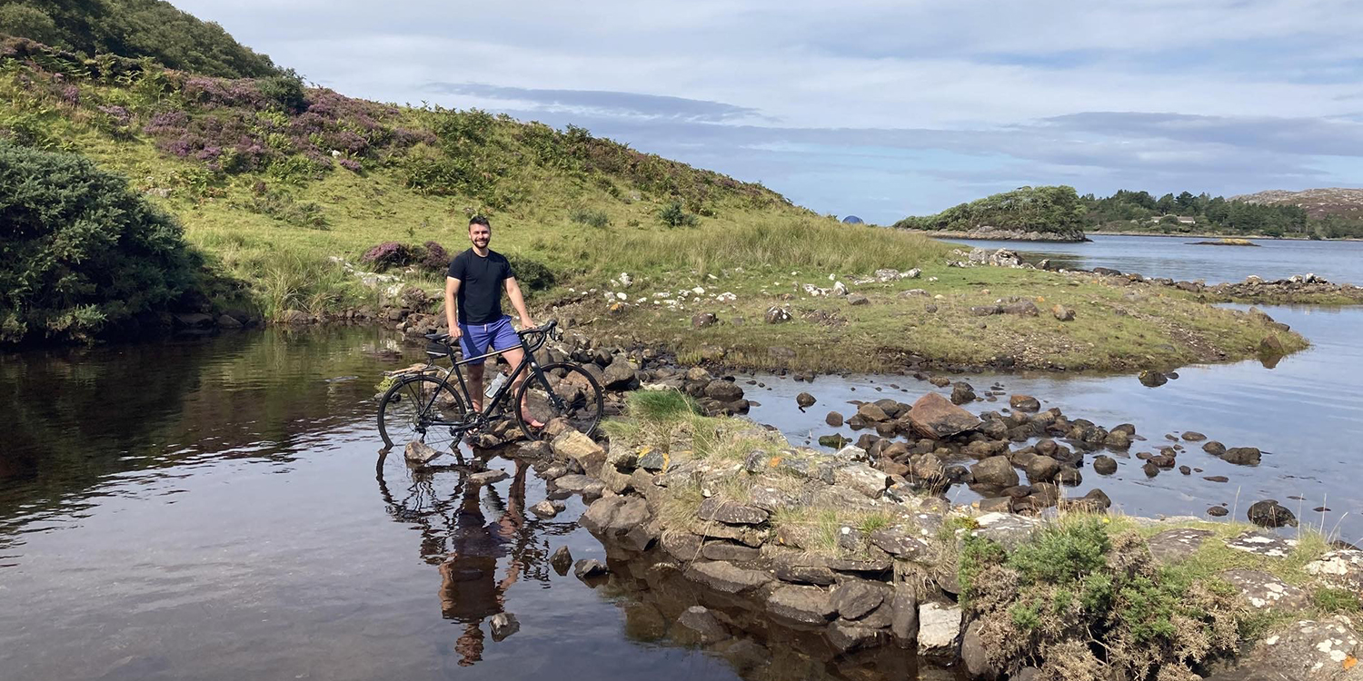 A man and his bike stand half in the water of a loch, a rough, rocky path leads across it.