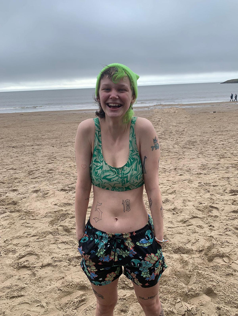 a person with green and black hair standing on a beach with the sea behind them. they are wearing a green bikini top and swimming trunks with aliens and flowers on them. they are smiling and have their hands in their pockets. they are wearing a bright green beanie.