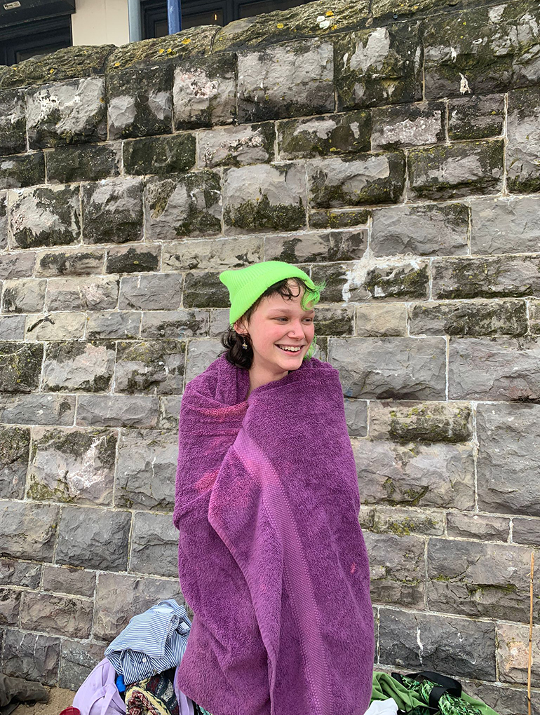 a person with green and black hir standing on a beach with a grey stone wall behind them smiling wrapped in a purple towel wearing a bright green hat 