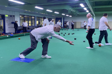 Man throwing a biased bowl towards the jack with other men playing bowls indoors behind him.