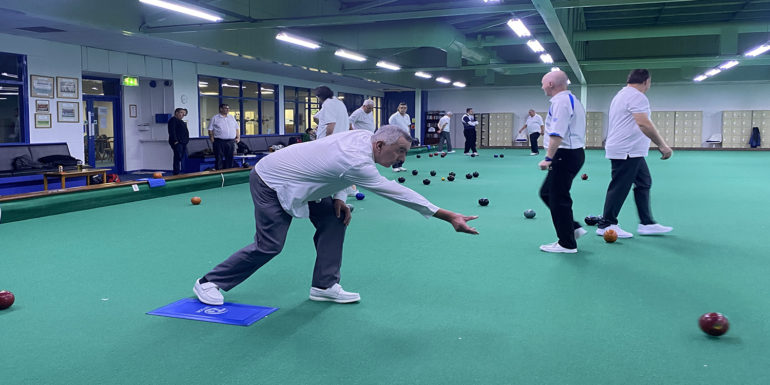 Man throwing a biased bowl towards the jack with other men playing bowls indoors behind him.