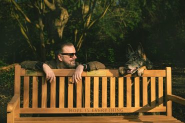 Ricky Gervais poses with a dog behind a bench.
