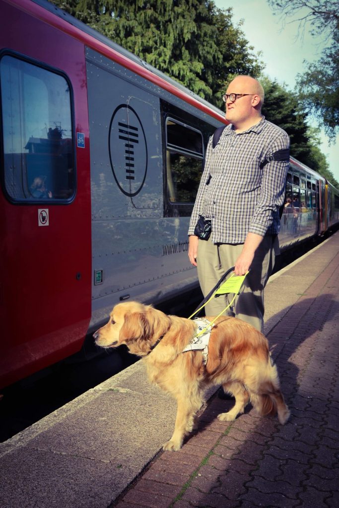 Photo of Ryan Moreland and his guide dog Jamie stood on a train station platform, looking out at the oncoming train.