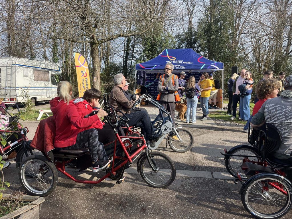 Two trikes, each with two people riding them, in front of Cereba's stall. There are people standing around in the background.